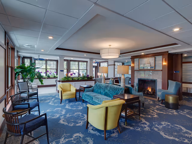 A cozy and well-lit living room area with a blue patterned carpet, a teal sofa, two yellow armchairs, and several dark wooden chairs. The room features a fireplace with a painting above it, two table lamps on side tables, large windows with plants on the windowsills, and a modern ceiling light fixture.