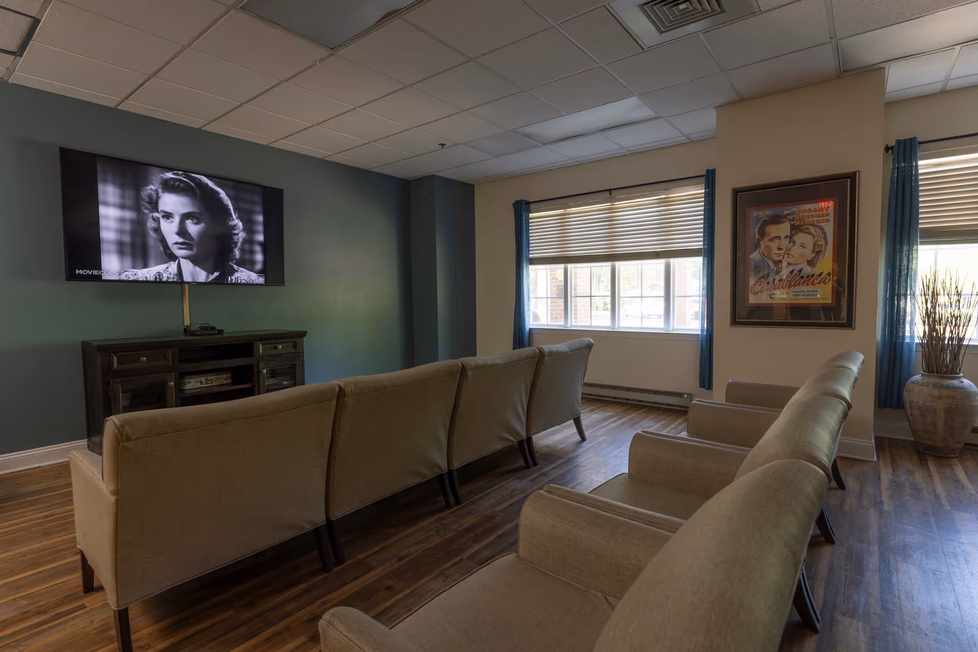 A cozy living room with two rows of beige armchairs facing a wall-mounted flat-screen TV displaying a black-and-white movie scene. The room has wooden flooring, a large window with blue curtains, a framed Casablanca movie poster on the wall, and a decorative vase with tall dried plants in the corner.