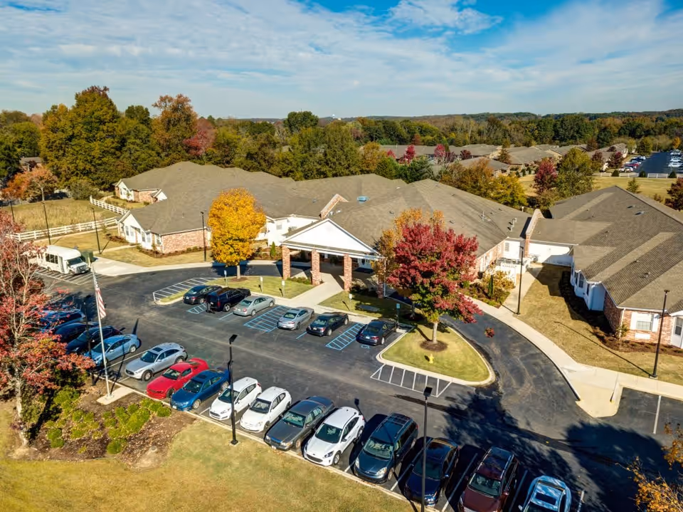 Aerial view of The Renaissance of Florence senior living facility showing multiple connected single-story buildings with gray roofs surrounded by trees with autumn foliage. There is a parking lot in front with several cars parked and a circular driveway leading to the main entrance with a covered portico. The area is landscaped with grass, trees, and shrubs under a partly cloudy sky.