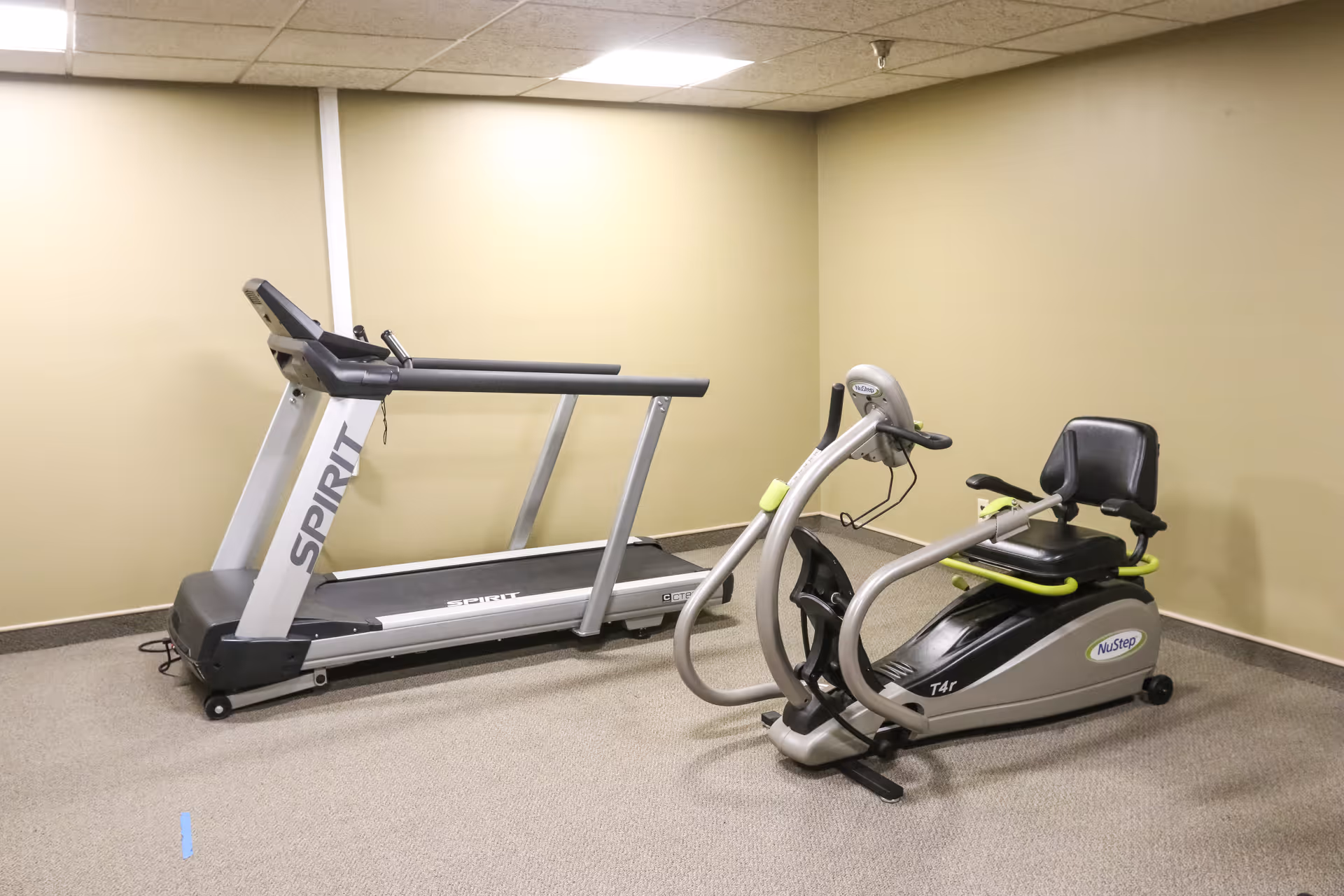A small exercise room with a treadmill and a NuStep T4r recumbent cross trainer on a carpeted floor, beige walls, and a drop ceiling.