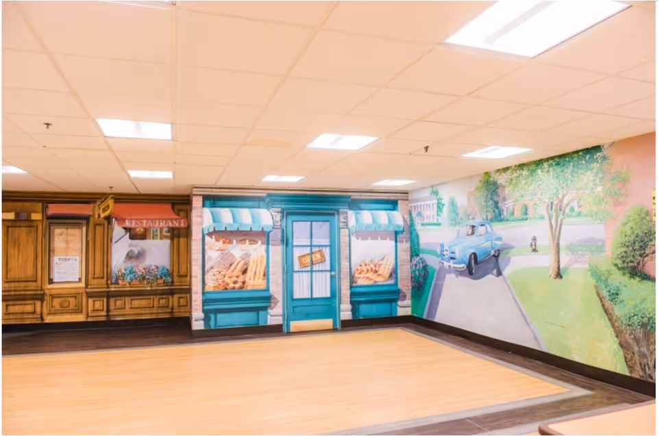 An interior room with walls painted to resemble a quaint street scene, including a bakery storefront with an 'Open' sign and a mural of a blue vintage car driving down a tree-lined road. The floor is wood-patterned, and the ceiling has recessed lighting.