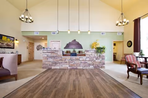 A spacious senior living facility lobby with a stone reception desk in the center, green accent wall behind it, and high vaulted ceilings with hanging light fixtures. There are chairs and a small table on the right side near a window with curtains, and a couch on the left side. The floor transitions from wood in the foreground to carpet near the reception area.