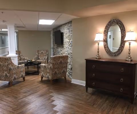 A cozy interior seating area with four patterned armchairs arranged around a small black coffee table. The room features wood-look flooring, a stone accent wall with a mounted flat-screen TV, and a wooden dresser with two lamps and an ornate oval mirror above it.
