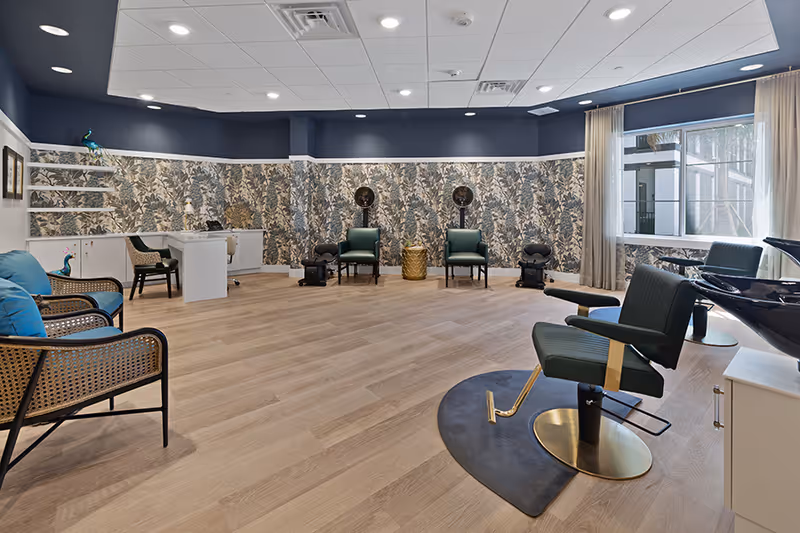 A spacious salon room with wooden flooring, featuring salon chairs, hair drying stations, and a seating area with blue cushions. The walls have a floral patterned wallpaper with a dark blue upper section. Large windows with sheer curtains allow natural light into the room.