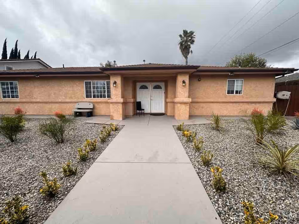 Front exterior view of a single-story building with a beige stucco finish, two white doors at the entrance, windows on either side, a concrete walkway leading up to the entrance, and landscaped areas with small plants and rocks on both sides. A palm tree and cloudy sky are visible in the background.