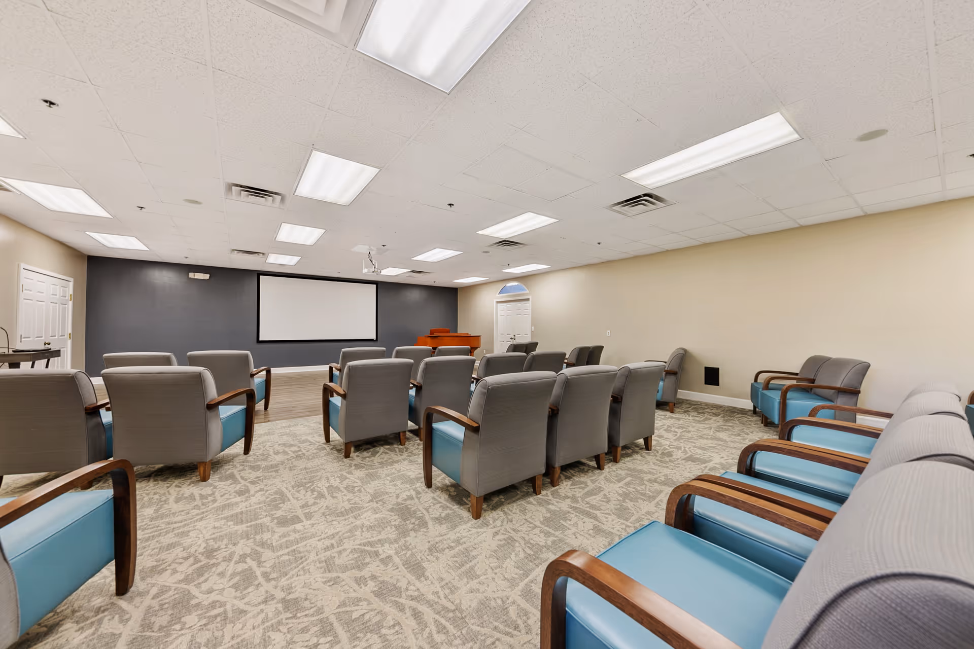 A large multipurpose meeting room with rows of upholstered armchairs facing a wall-mounted projection screen and a piano.