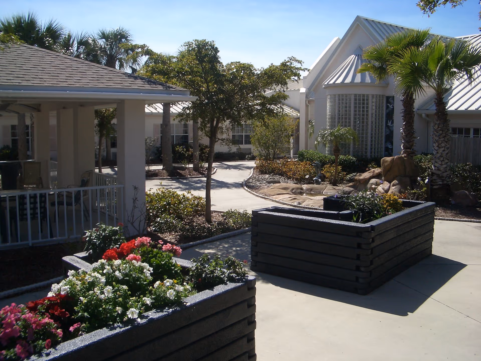 Outdoor courtyard area of a senior living facility with raised flower beds filled with colorful flowers, palm trees, a paved walkway, and white buildings with metal roofs in the background under a clear blue sky.