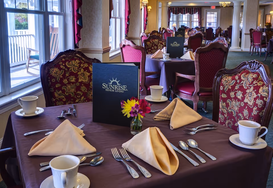Dining room table set with folded napkins, cups, silverware, a small vase of flowers and a Sunrise Senior Living menu in a formal senior-living dining room.
