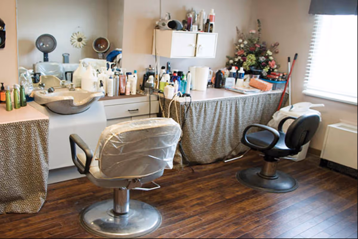 Interior of a hair salon area in a senior living facility with two salon chairs, a sink for washing hair, a large mirror, and various hair care products on the counter and shelves. There is a window with blinds letting in natural light and a floral arrangement on the counter.
