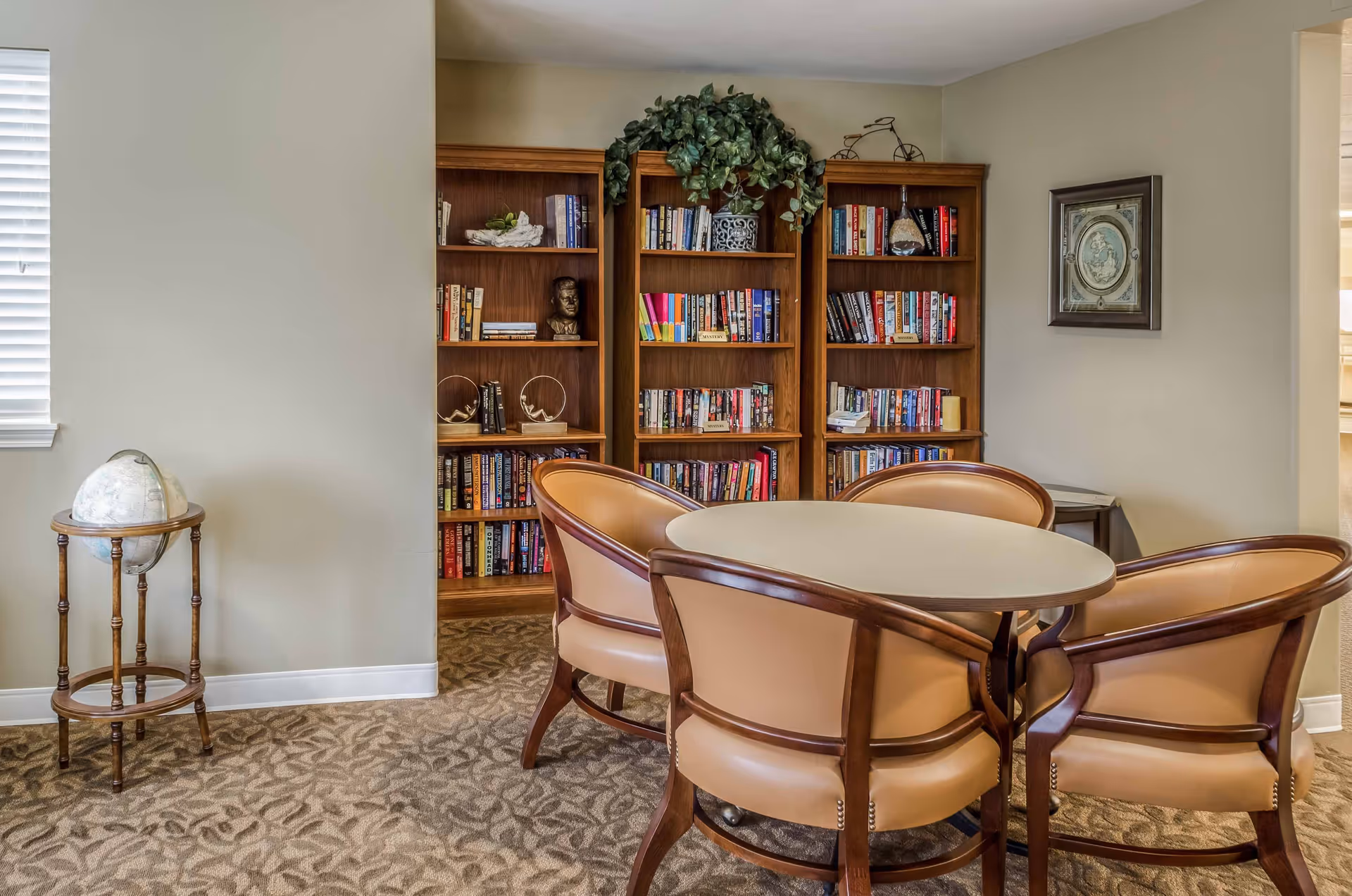 A cozy reading or meeting area in a senior living facility featuring a round table surrounded by four cushioned wooden chairs. Behind the table are three wooden bookshelves filled with books and decorative items, including a potted plant on top. To the left, there is a globe on a wooden stand near a window with blinds. A framed picture hangs on the wall to the right.