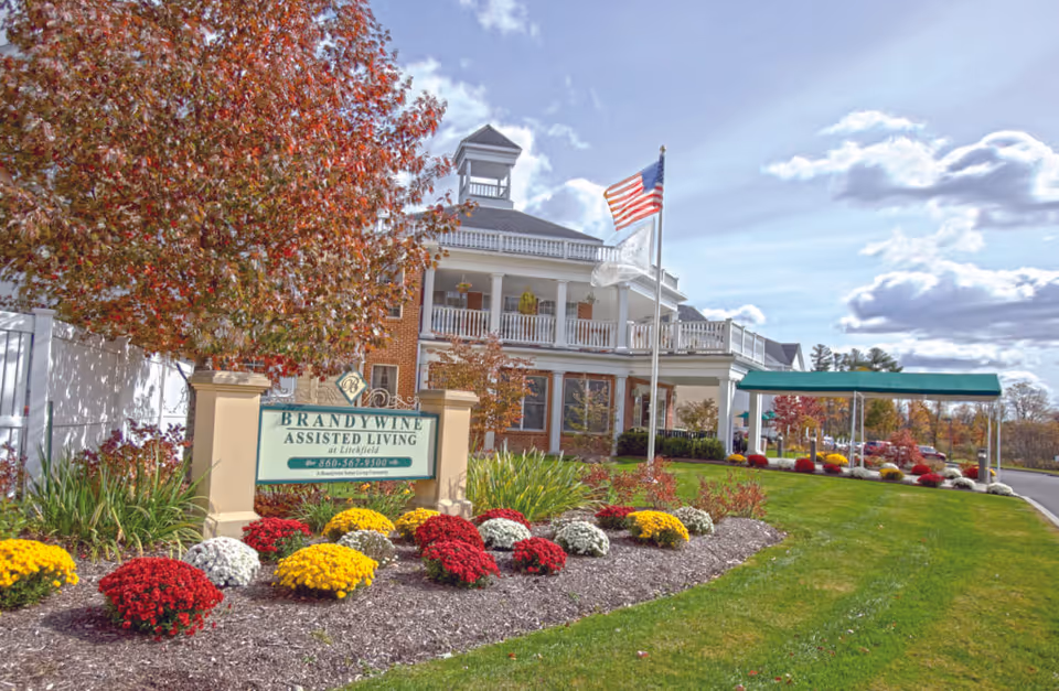 Front exterior view of Brandywine Assisted Living at Litchfield building with a landscaped garden featuring colorful flowers and a green lawn. An American flag flies on a flagpole near the entrance, and a green canopy covers the driveway.