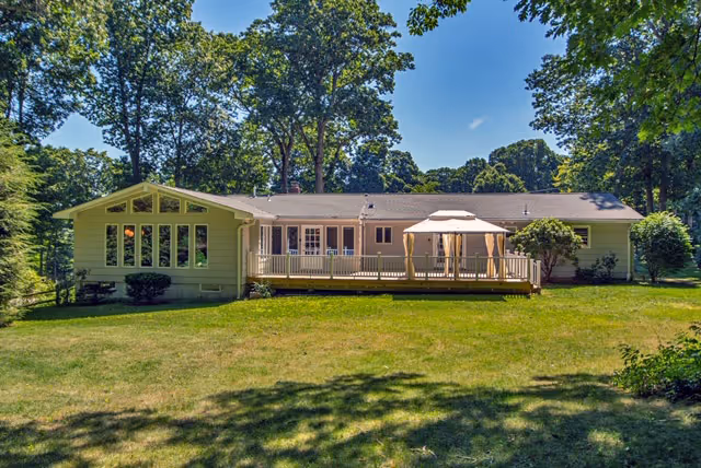 Single-story ranch-style house with a wooden deck and gazebo overlooking a grassy yard surrounded by trees.