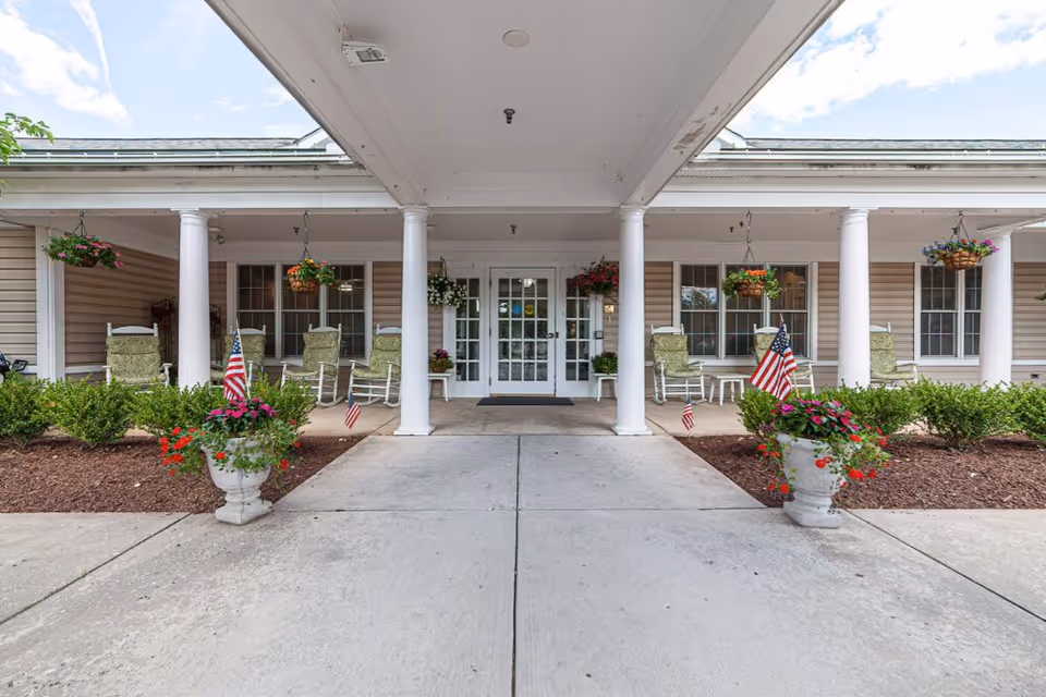 Entrance of HeartFields Assisted Living at Easton featuring a covered walkway with white columns, hanging flower baskets, rocking chairs with green cushions, American flags in flower pots, and glass double doors leading inside.