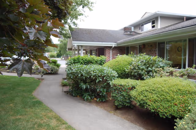 Side view of a single-story brick building with large windows, surrounded by well-maintained bushes and greenery along a concrete walkway under a partly cloudy sky.