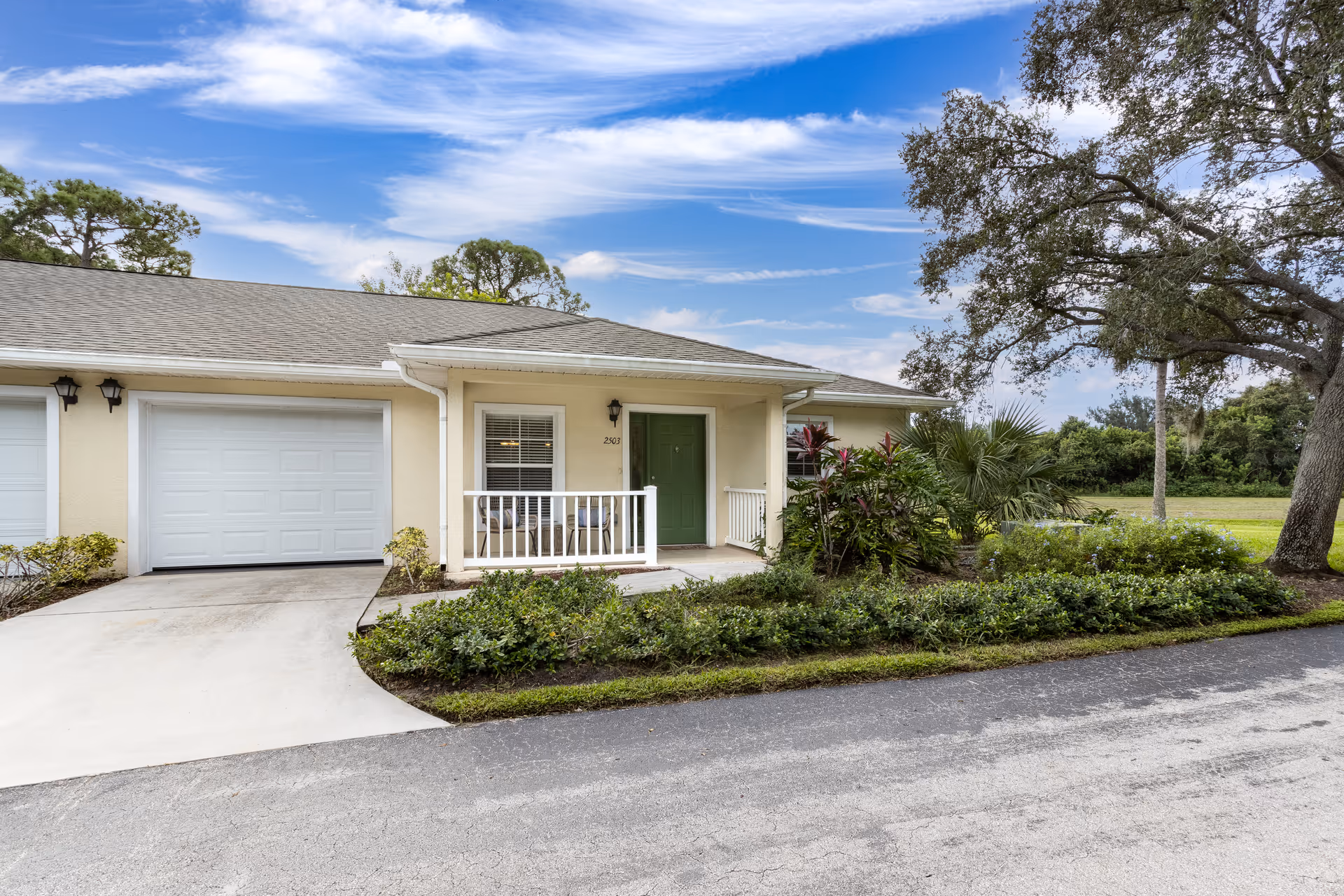 Single-story beige senior living unit with a garage, small porch with a green door and landscaping under a partly cloudy sky.