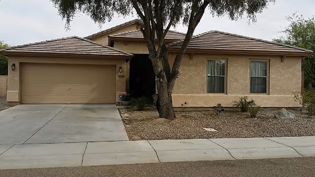 Front exterior view of a single-story beige stucco house with a two-car garage, a tree in the front yard, and a gravel landscape with some small plants.