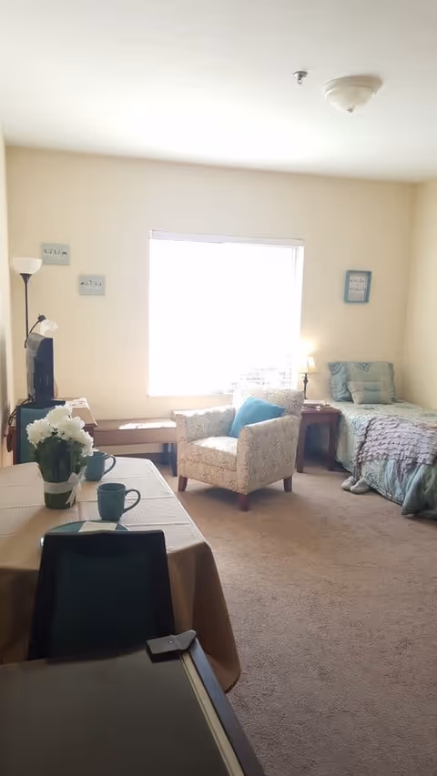 Bright assisted-living studio room with a bed, armchair, table set with mugs and flowers, a TV, and a large window.
