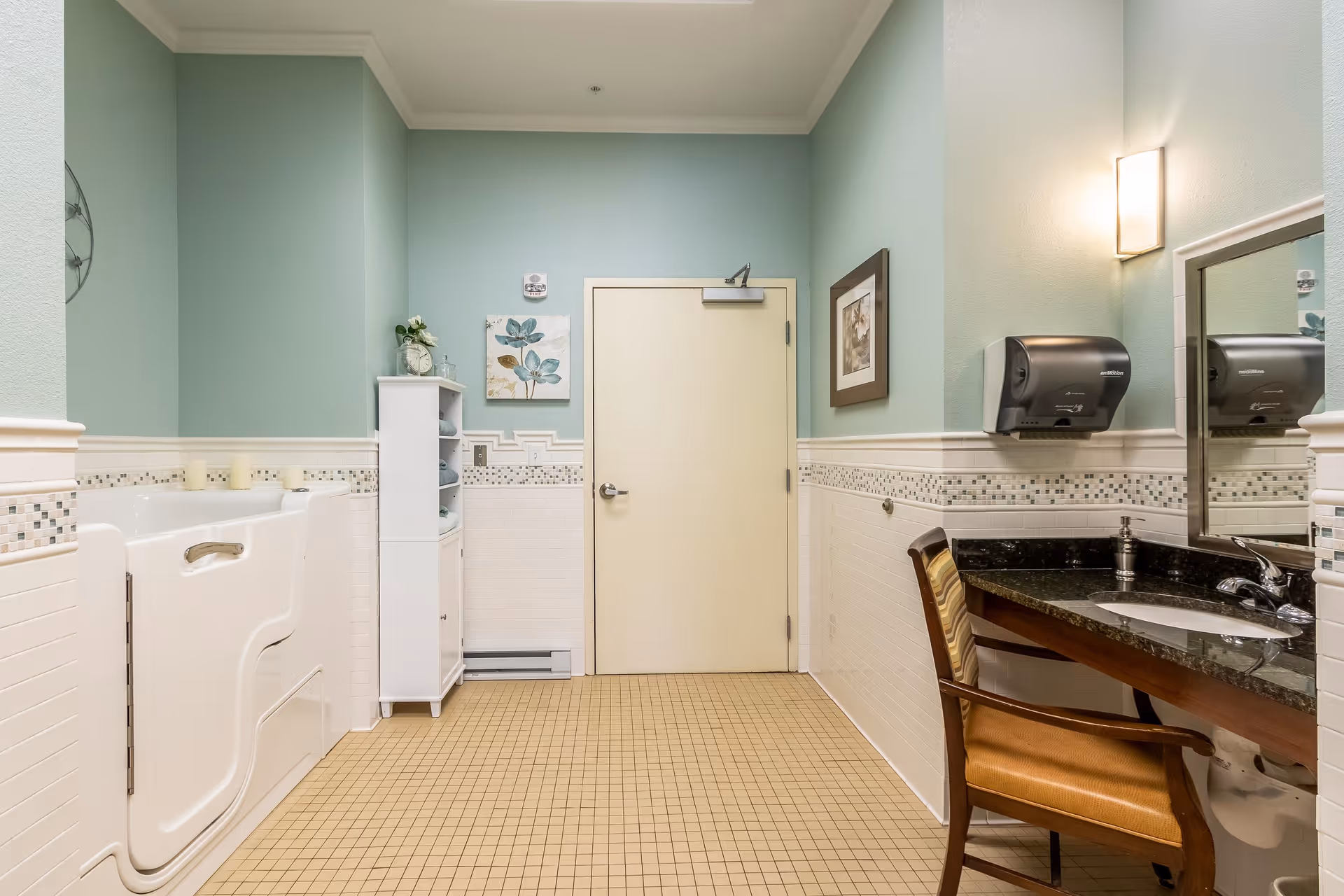 A clean and accessible bathroom with light green walls and beige tiled floor. The room features a walk-in bathtub with grab bars on the left, a white storage cabinet with towels and decorative items, a sink with a granite countertop and a wooden chair on the right, a wall-mounted paper towel dispenser, a mirror, and framed floral artwork on the walls.