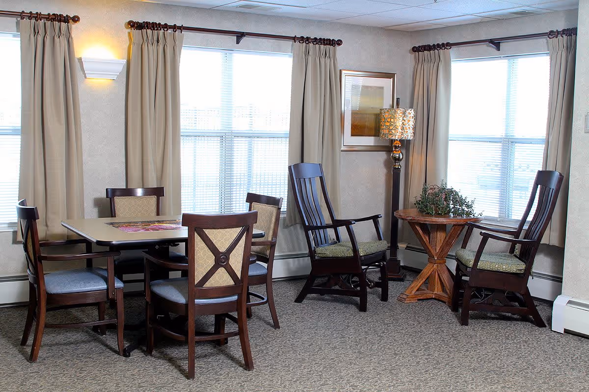 A cozy senior living common area with a rectangular dining table surrounded by four wooden chairs with cushioned seats. Nearby, there are two wooden rocking chairs with cushions around a small round wooden table holding a green plant. The room has beige walls, carpeted floor, large windows with beige curtains, a floor lamp with a patterned shade, and a framed picture on the wall.