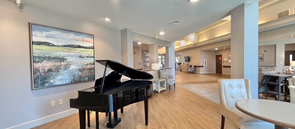 A bright senior living common area with a black grand piano, seating, and a large landscape painting on the wall.