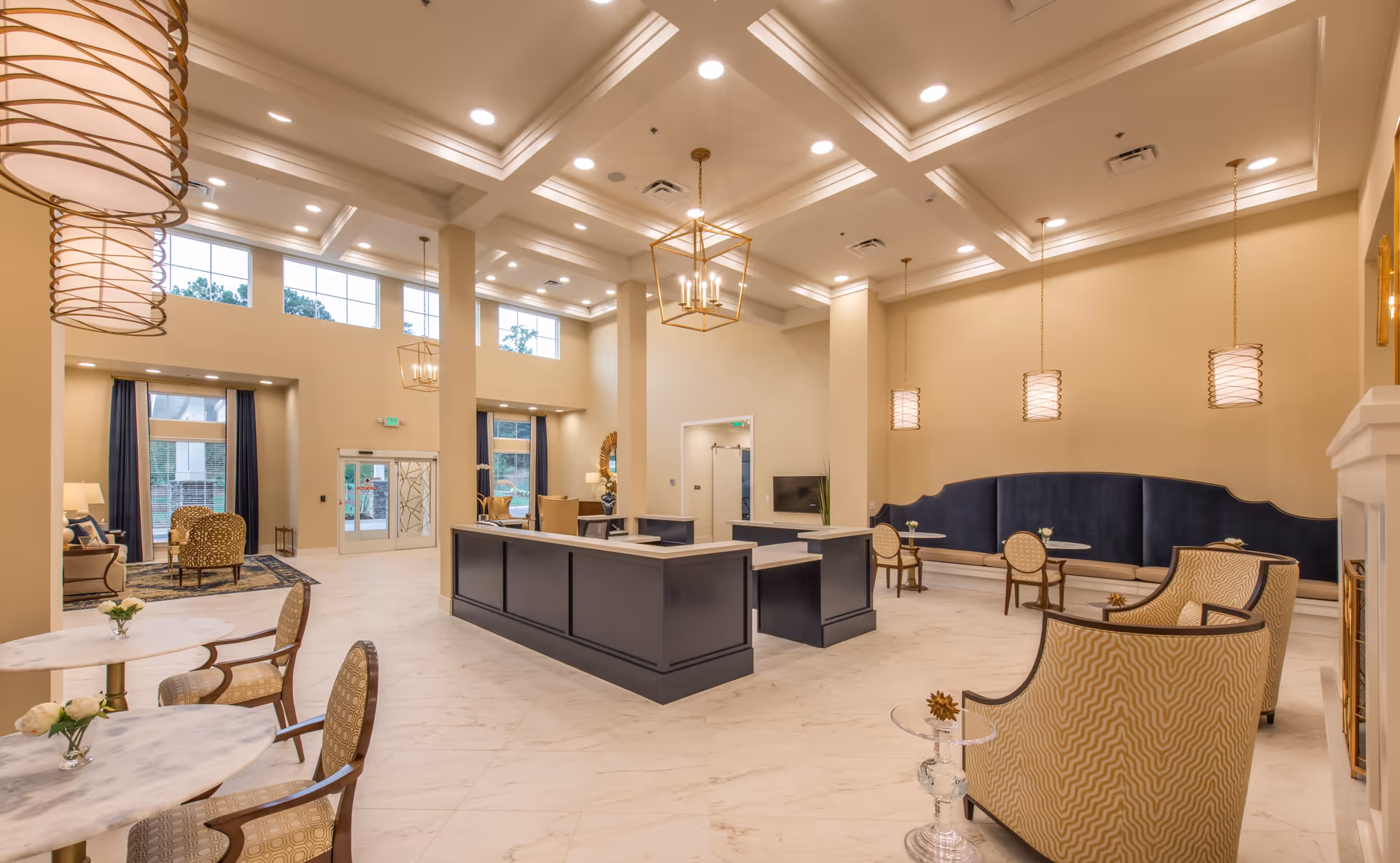 A spacious and elegant senior living facility lobby with high ceilings, large windows, and modern lighting fixtures. The room features a central reception desk in dark blue, several seating areas with upholstered chairs and small round tables, and a long blue cushioned bench along one wall. The floor is light-colored marble, and the walls are painted in a soft beige tone. There are decorative elements such as flowers on tables and a fireplace on the right side.