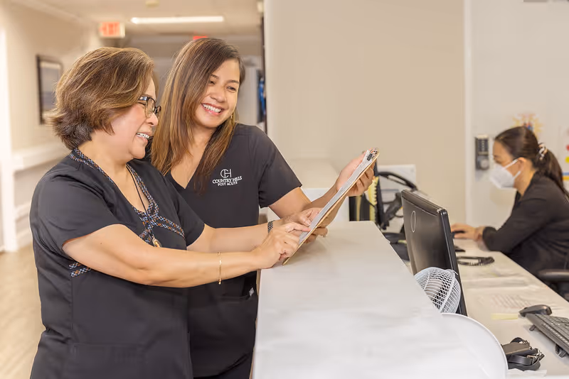 Two healthcare workers in black scrubs smiling and discussing notes on a clipboard at a reception desk, with a third person working on a computer in the background wearing a face mask.