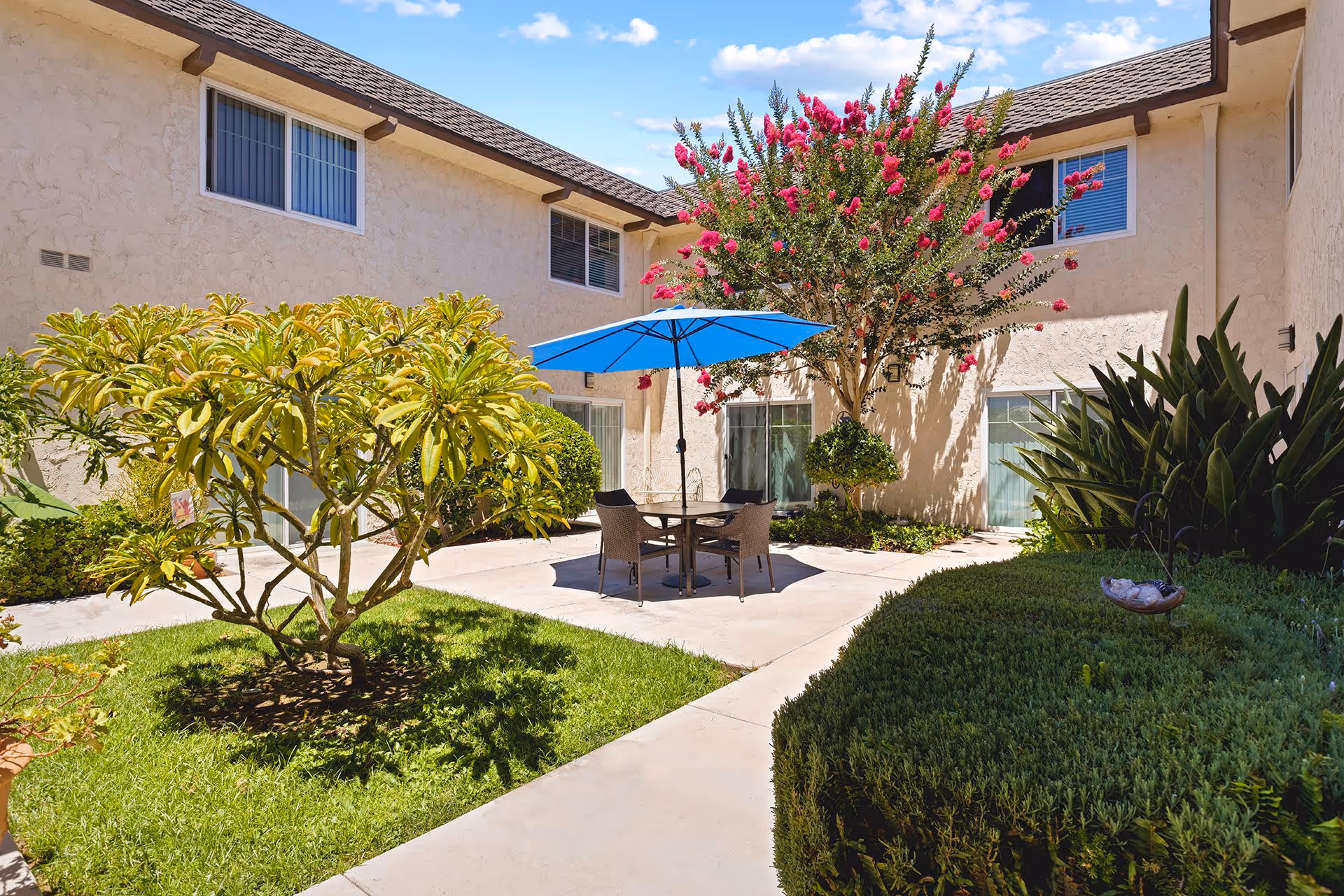 Sunny courtyard with a central patio table and blue umbrella surrounded by landscaped shrubs and a two-story beige building.