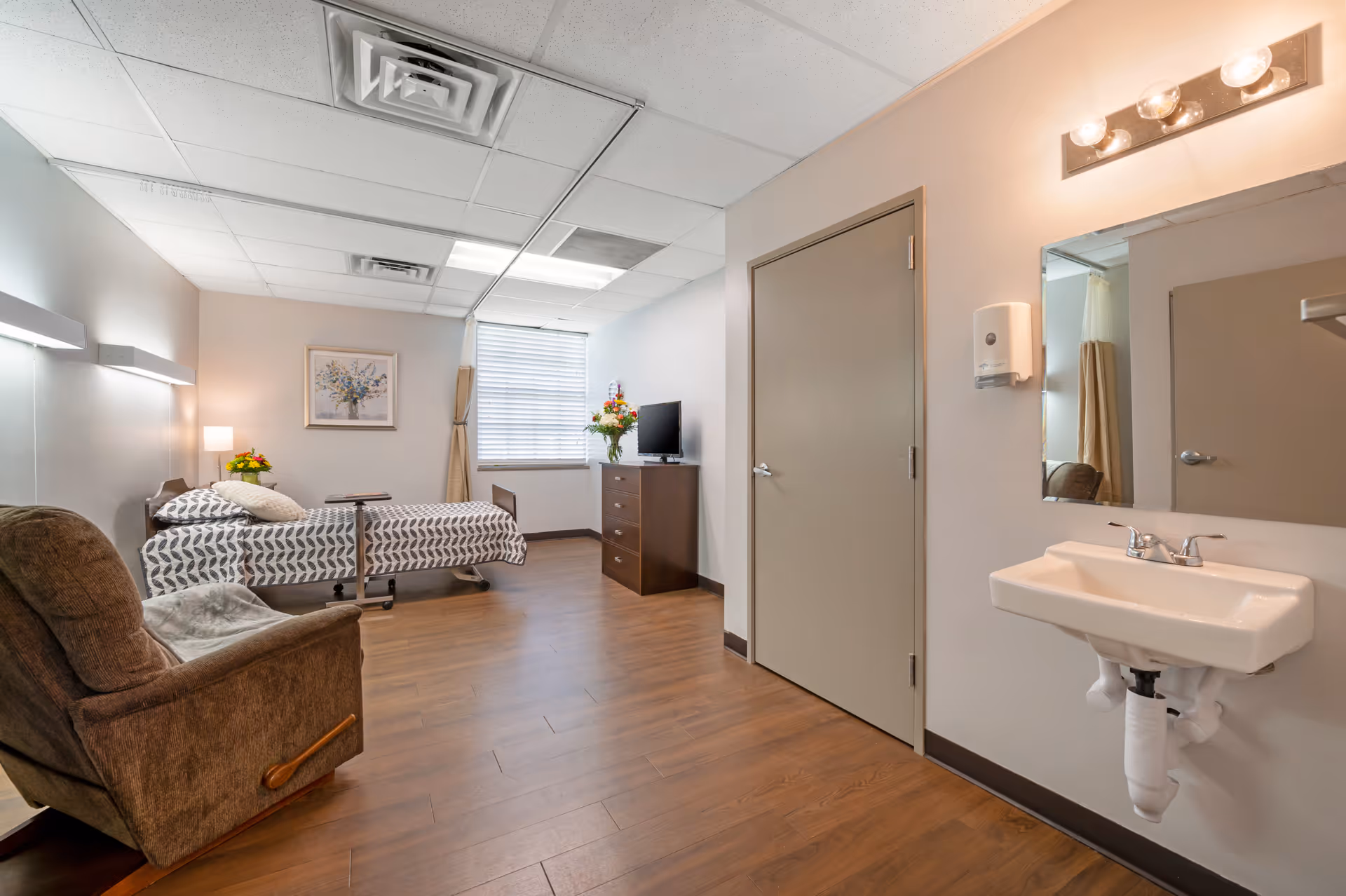 A clean and simple bedroom in a rehabilitation and health care center featuring a single bed with patterned bedding, a brown recliner chair, a wooden dresser with a flat-screen TV and a vase of flowers, a window with blinds and curtains, a wall-mounted sink with a mirror and light fixtures above, and a closed door. The room has wood flooring and neutral-colored walls.
