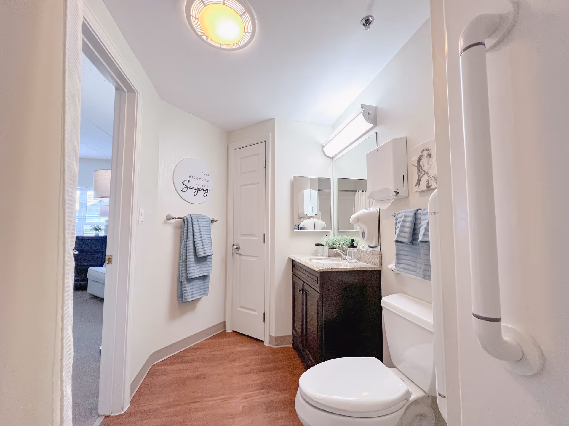 Bright and clean senior living bathroom with a white toilet, dark wood vanity with a sink, a large mirror, blue towels hanging on a rack, a grab bar near the toilet, and a door leading to another room with a window and lamp visible.