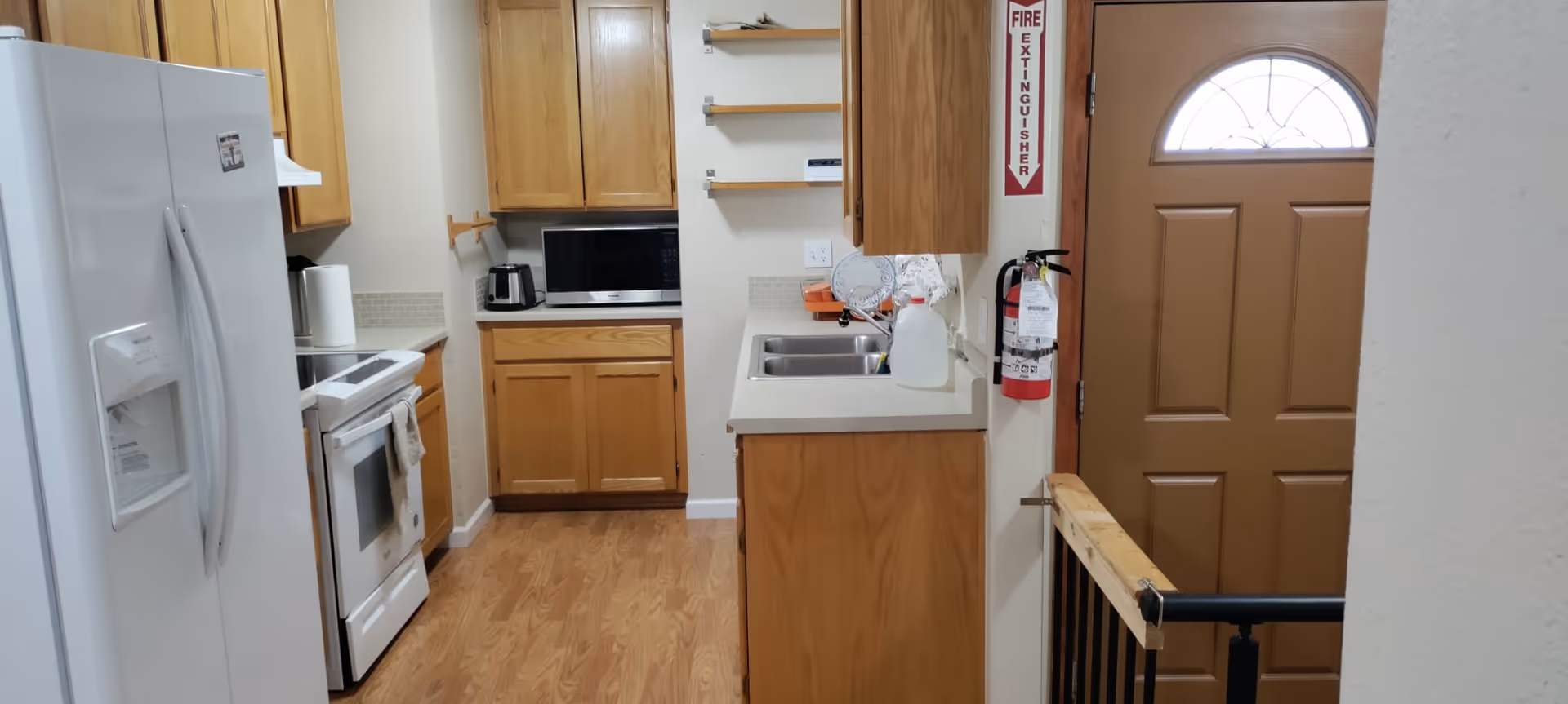 A small kitchen area with wooden cabinets, a white refrigerator, a white stove with an oven, a microwave on the countertop, a double sink, and a brown front door with a window at the top. There is a fire extinguisher mounted on the wall near the door and a wooden floor.