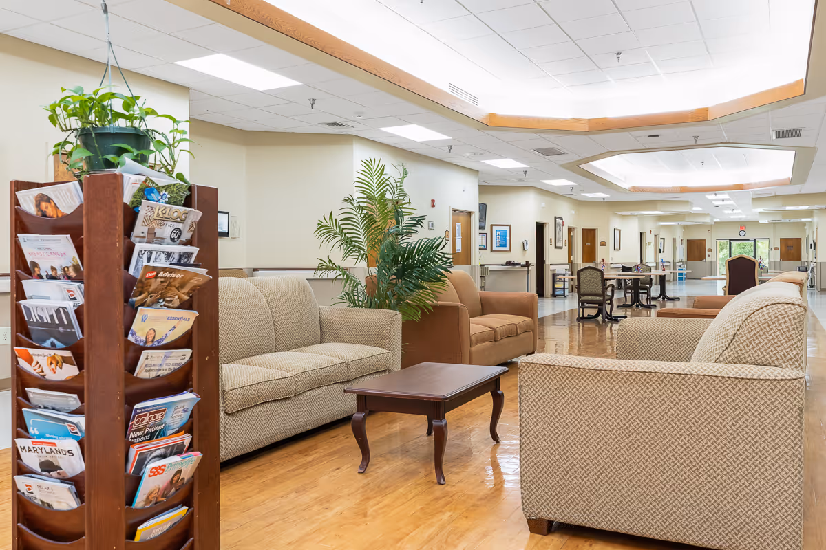 A spacious and well-lit common area in a senior living facility featuring beige and brown upholstered sofas and armchairs arranged around a wooden coffee table. A wooden magazine rack filled with various magazines and brochures is positioned on the left side. The room has a polished wooden floor, potted plants, and a high ceiling with recessed lighting. In the background, there are additional tables and chairs along a wide hallway with doors and framed pictures on the walls.