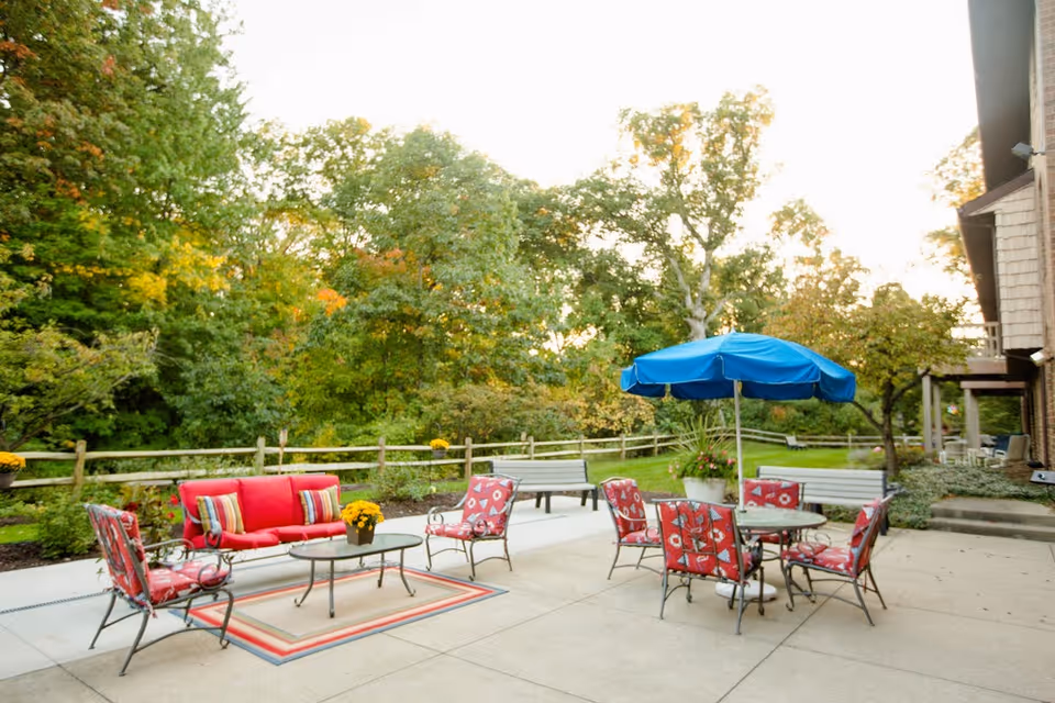 Outdoor patio area with red cushioned chairs and a sofa around a glass coffee table on a rug, a round table with red cushioned chairs and a blue umbrella, surrounded by green trees and a wooden fence.