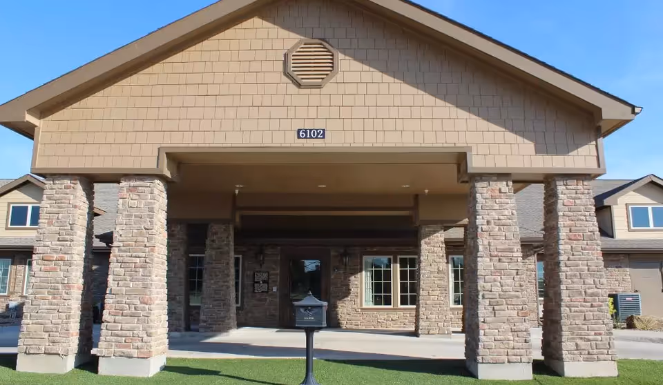 Front exterior view of a building with a covered entrance supported by large stone pillars. The building has beige siding and a sign with the number 6102 above the entrance. The sky is clear and blue.