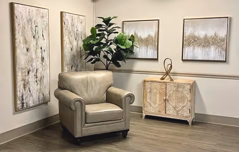 A leather armchair sits in a corner next to a decorative cabinet, potted plant, and three framed abstract paintings on a wood floor.