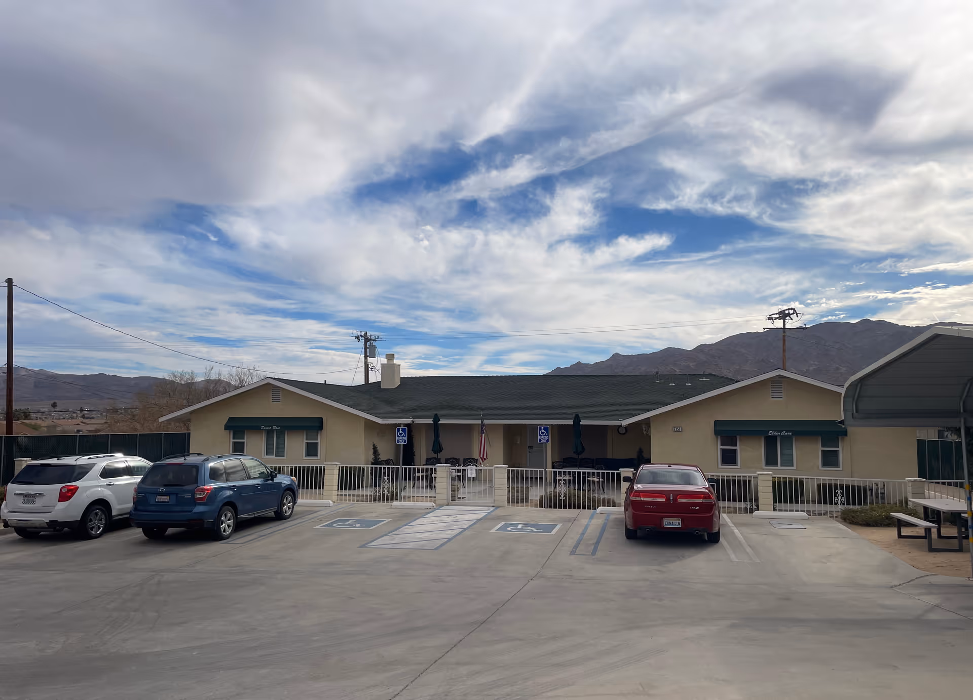 Front exterior view of Desert Rose Elder Care, Inc. building with a parking lot in front, including three parked cars and three handicap parking spaces. The building is a single-story structure with a green roof and beige walls, set against a backdrop of mountains and a partly cloudy sky.