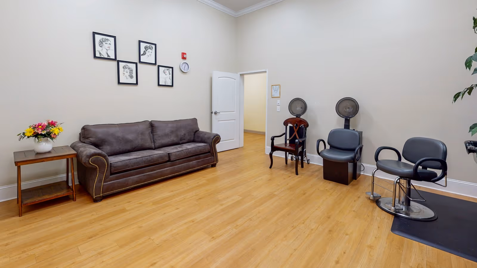 A spacious room with light wood flooring and beige walls featuring a brown leather sofa with nailhead trim, a small wooden side table with a vase of colorful flowers, four framed black and white portraits on the wall, and three salon chairs with hair dryers attached. An open white door leads to another room.