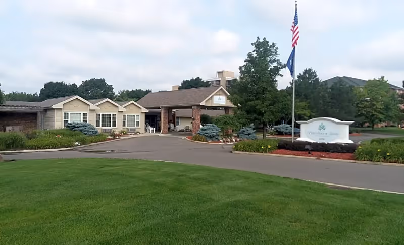 Front entrance of a senior living facility with a covered drop-off, American flag, sign, and landscaped lawns.