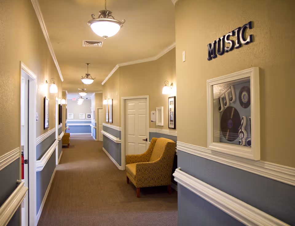 Well-lit carpeted hallway in a senior living facility with chairs, framed artwork and a 'MUSIC' sign on the wall.