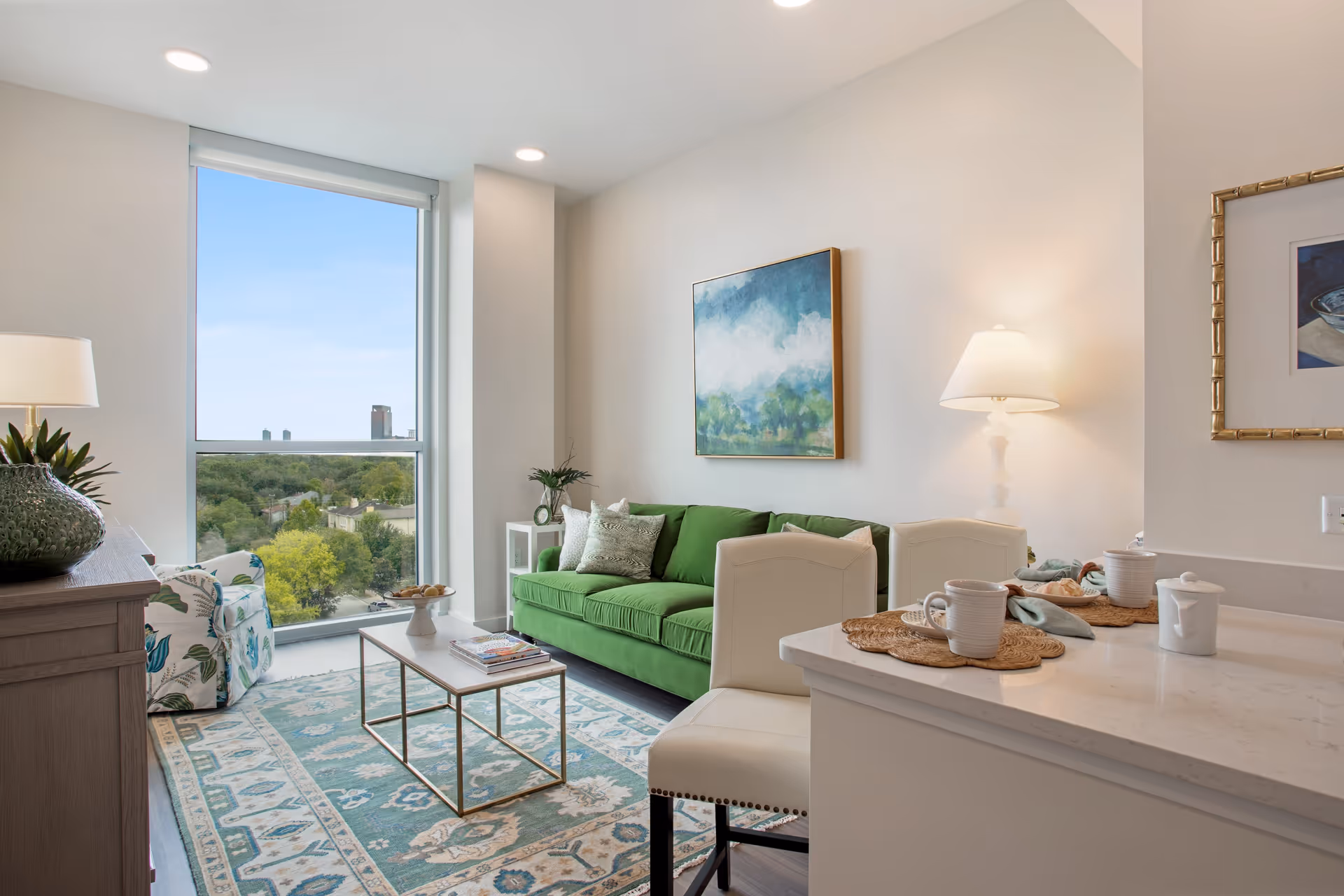 A bright and modern living room with a large window offering a view of trees and buildings outside. The room features a green sofa with decorative pillows, a patterned area rug, a coffee table with magazines, a floral armchair, a wooden sideboard with a plant, and a countertop with two white chairs, cups, and a lamp on the wall.