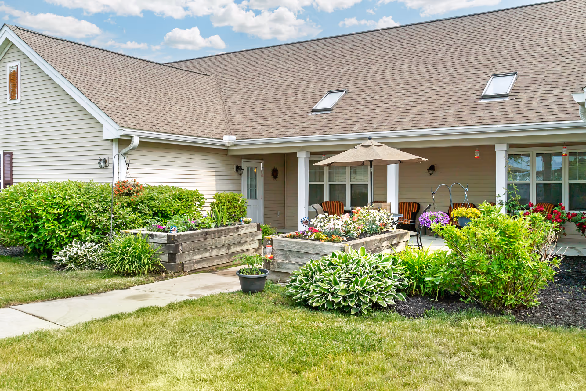 Outdoor patio area of a senior living facility with raised wooden garden beds filled with colorful flowers, green shrubs, and a small potted plant. The building has beige siding, a brown shingled roof with skylights, and a covered porch with chairs and a table under a large umbrella. The sky is partly cloudy.