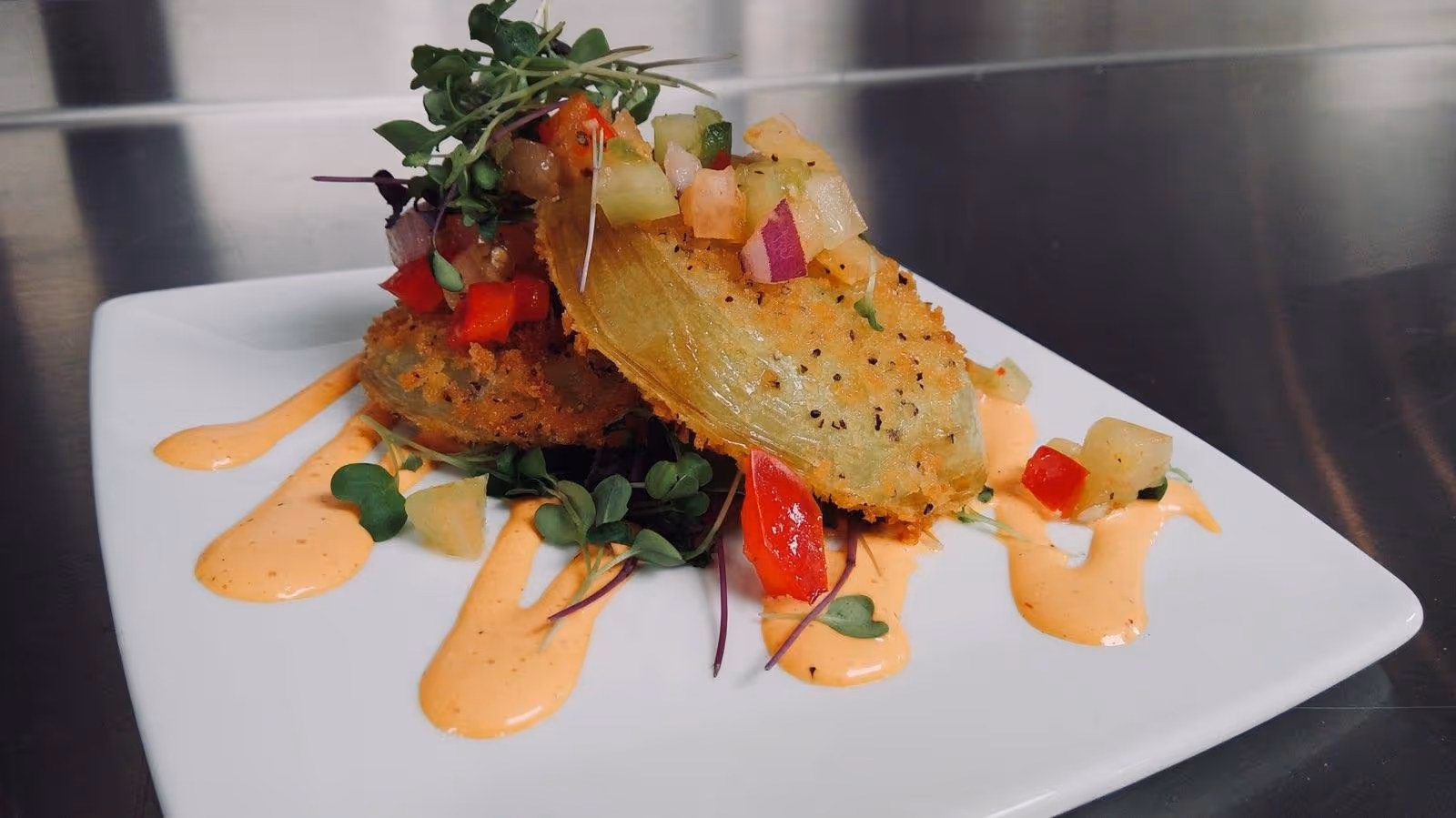 A gourmet dish featuring two breaded and fried artichoke hearts topped with diced vegetables and microgreens, served on a white plate with drizzles of a creamy orange sauce.