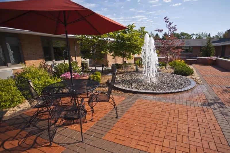 Outdoor patio area with a round metal table and four chairs under a large red umbrella, surrounded by brick paving. In the background, there is a circular water fountain with water spraying upwards, some greenery, and a brick building under a partly cloudy sky.