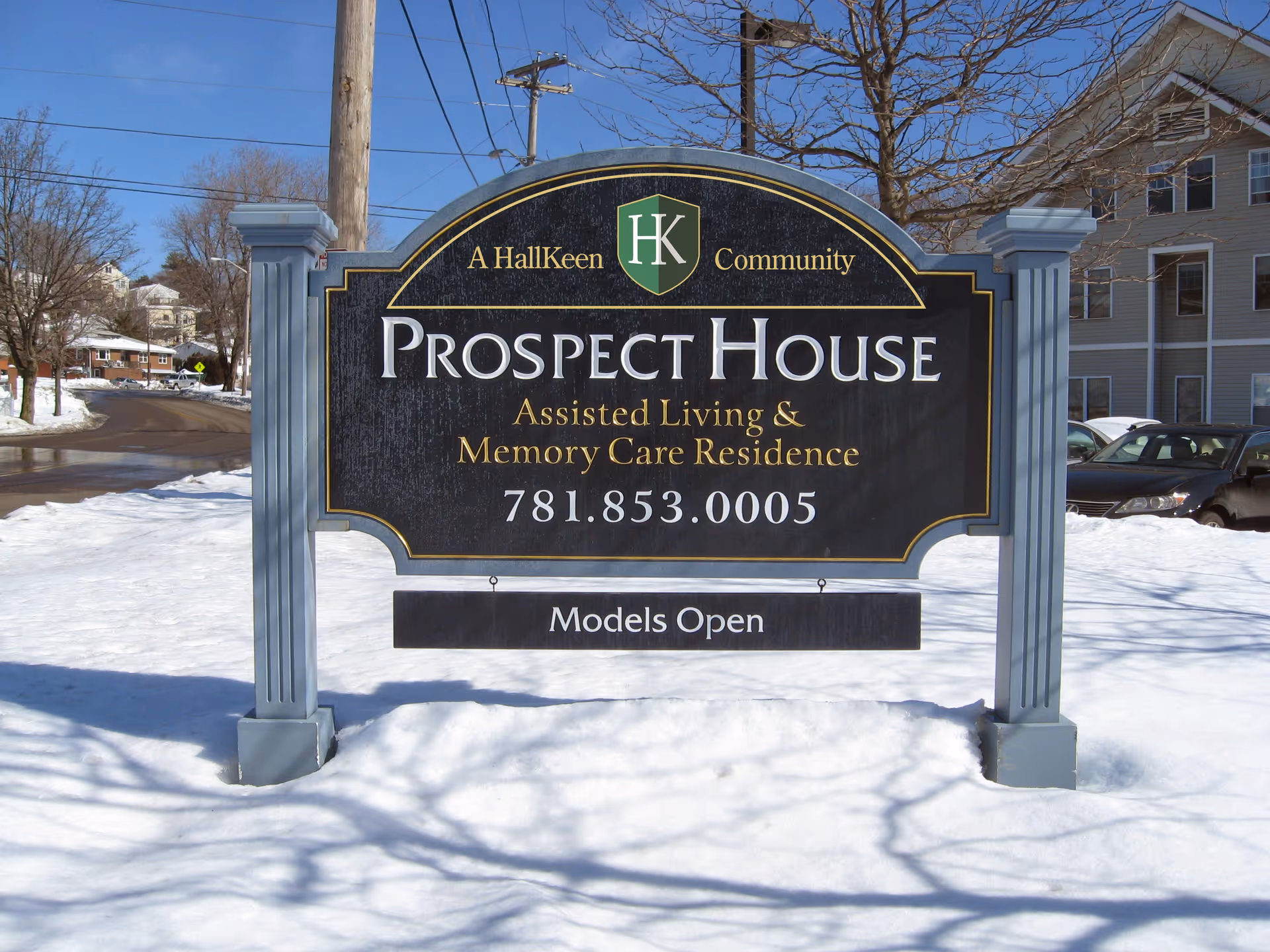 Outdoor sign for Prospect House Assisted Living and Memory Care Residence, mounted on two gray posts, with snow on the ground and a street and buildings in the background.