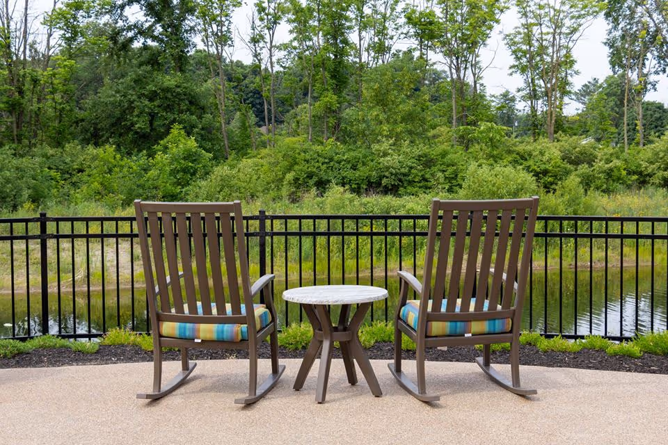 Two wooden rocking chairs with colorful cushions flank a small round table on a patio overlooking a pond and tree-lined greenery.