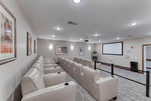 Interior view of a small theater room with two rows of beige leather recliner chairs facing a large white projection screen. The room has light gray walls adorned with framed artwork and is well-lit with ceiling lights. There is a projector mounted on the ceiling and a small railing separating the seating area from the screen area.