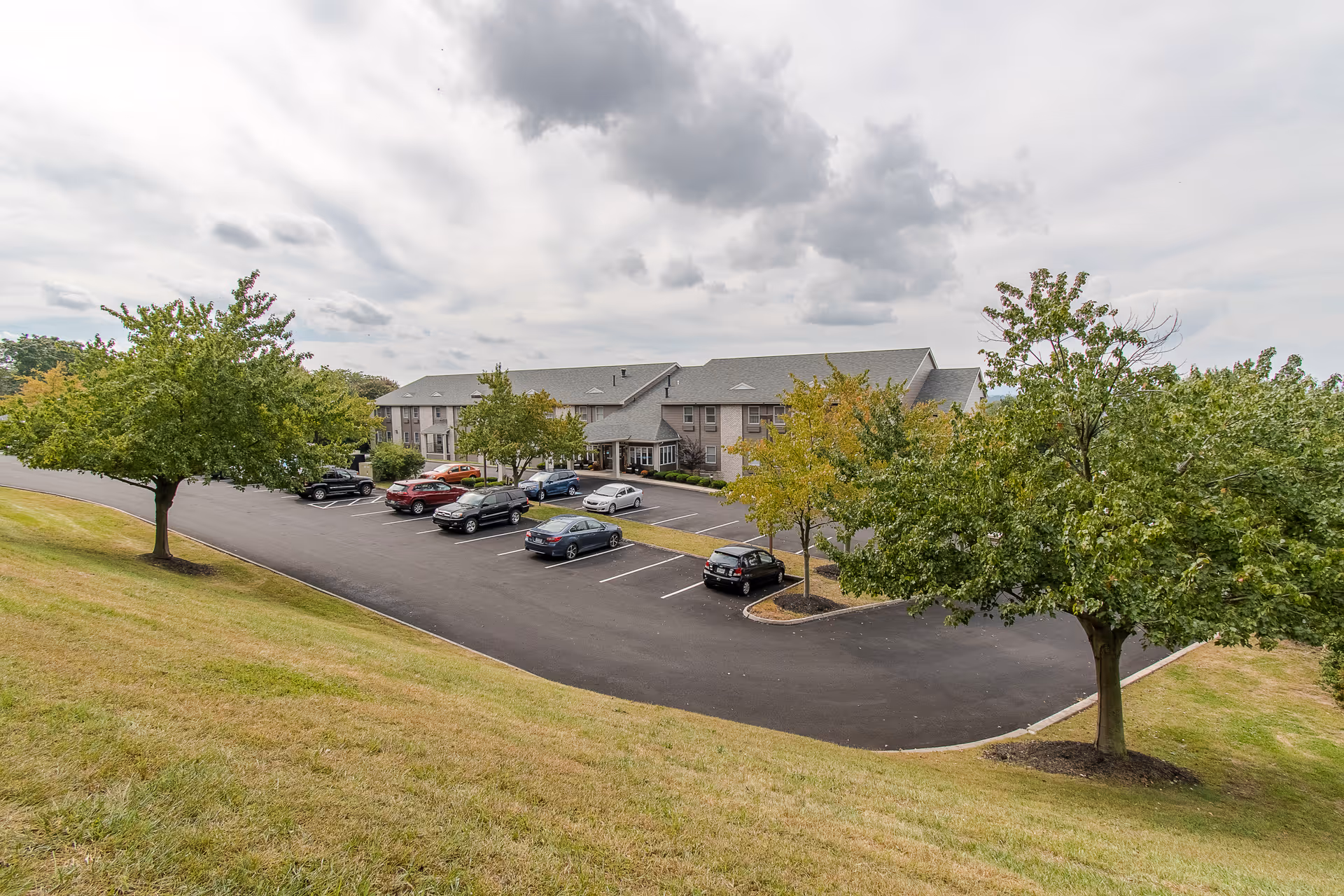 Exterior view of a senior living building with a parking lot, parked cars, trees on a grassy slope, and a cloudy sky.