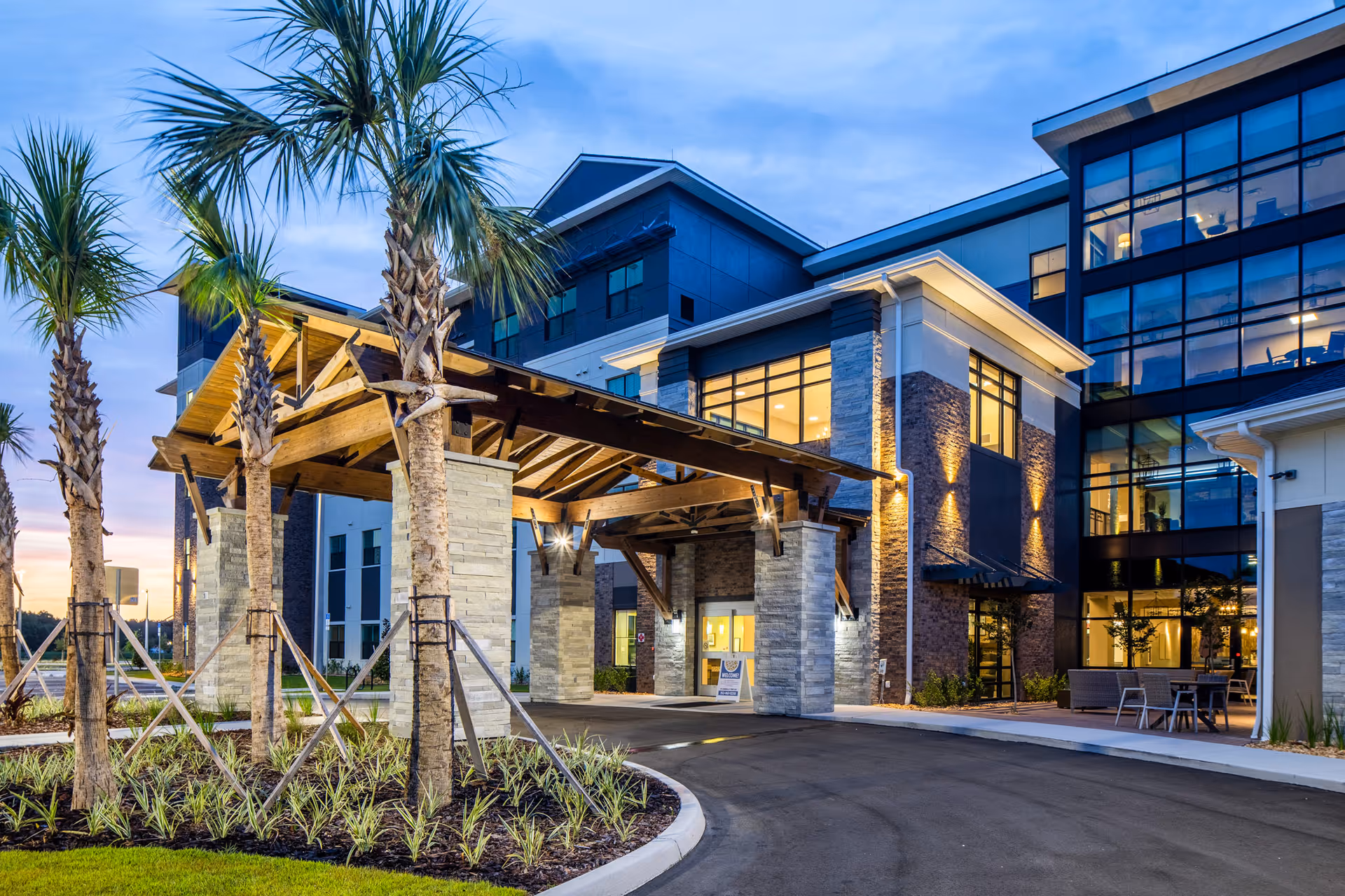 Front entrance of Blue Heron Senior Living with a wooden covered porte-cochère, palm trees, and illuminated glass façade at dusk.