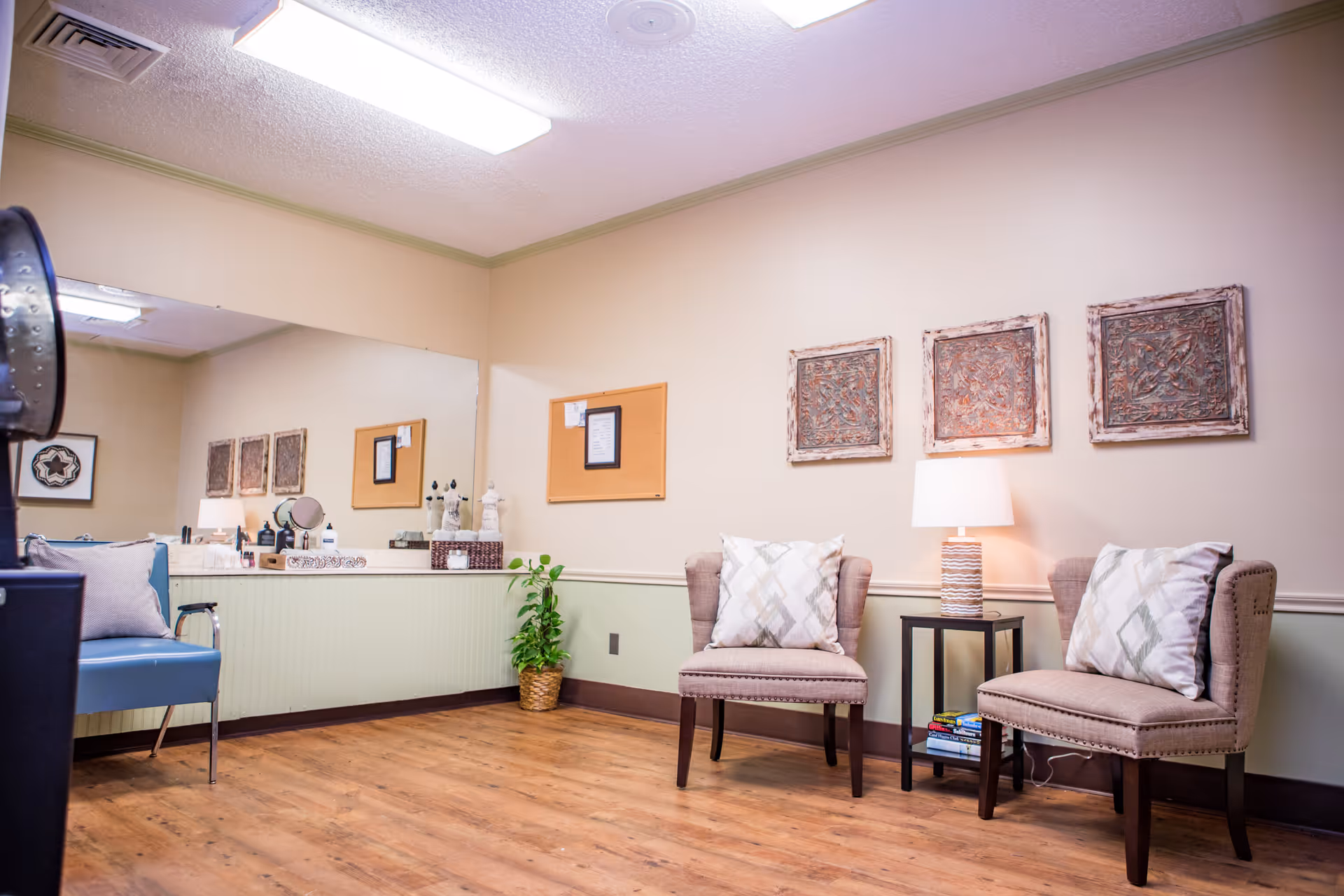 A cozy waiting or sitting area with two beige upholstered chairs with patterned pillows, a small black side table with a lamp and books, a large mirror on the wall, a blue chair, a plant in a woven basket, and decorative wall art. The room has wood flooring and soft lighting.