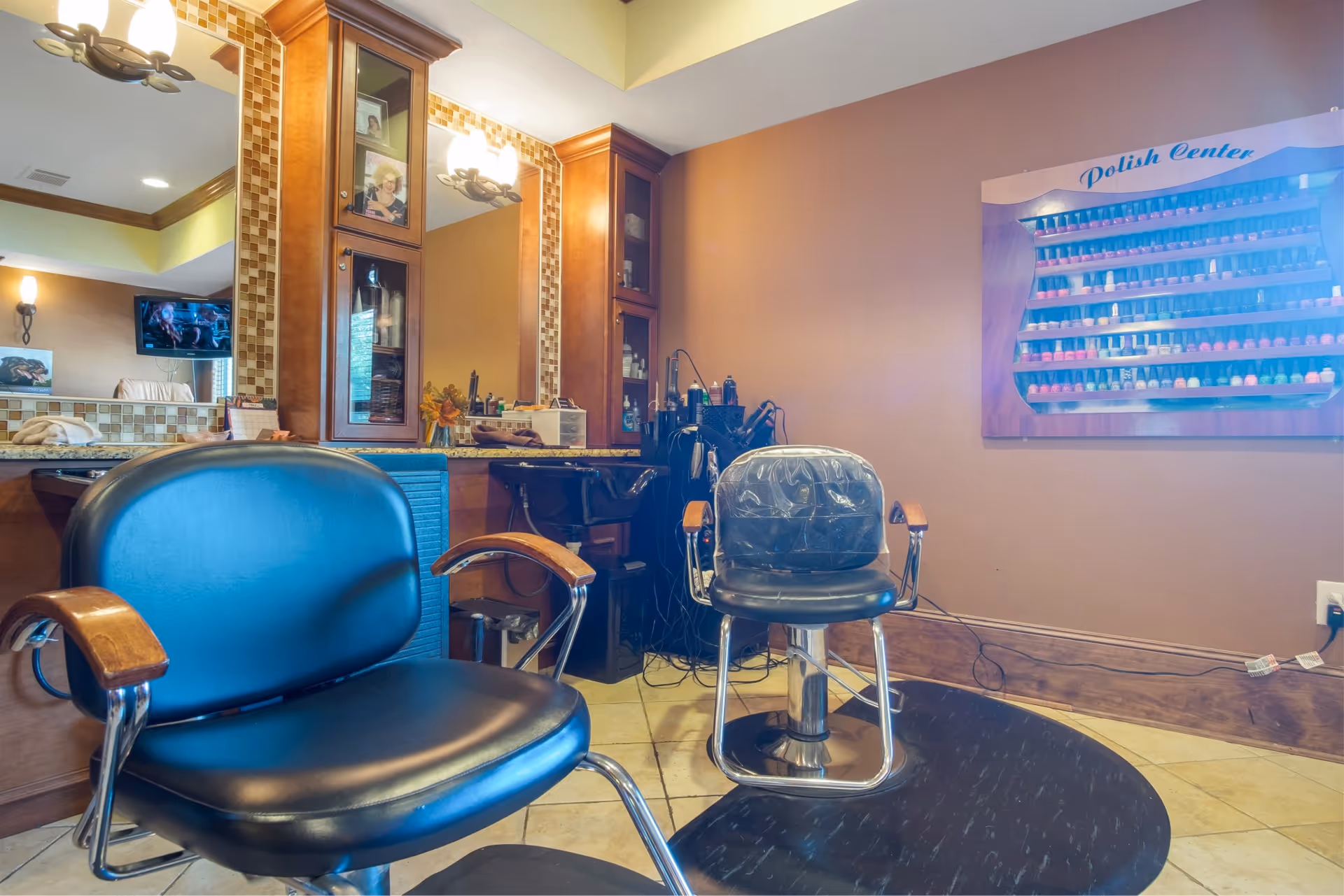 Interior view of a salon area with two black salon chairs with wooden armrests, a large mirror with wooden cabinets on either side, and a wall-mounted nail polish display labeled 'Polish Center'. The room has warm lighting and beige tiled flooring.