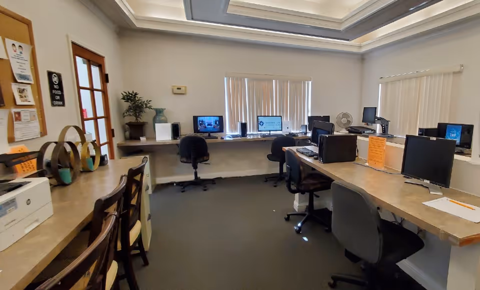 An office room with multiple computer workstations along the walls. There are several chairs, desktop computers, and laptops on the desks. The room has a bulletin board with notices on the left wall and vertical blinds covering the windows. The ceiling has recessed lighting with a tray ceiling design.